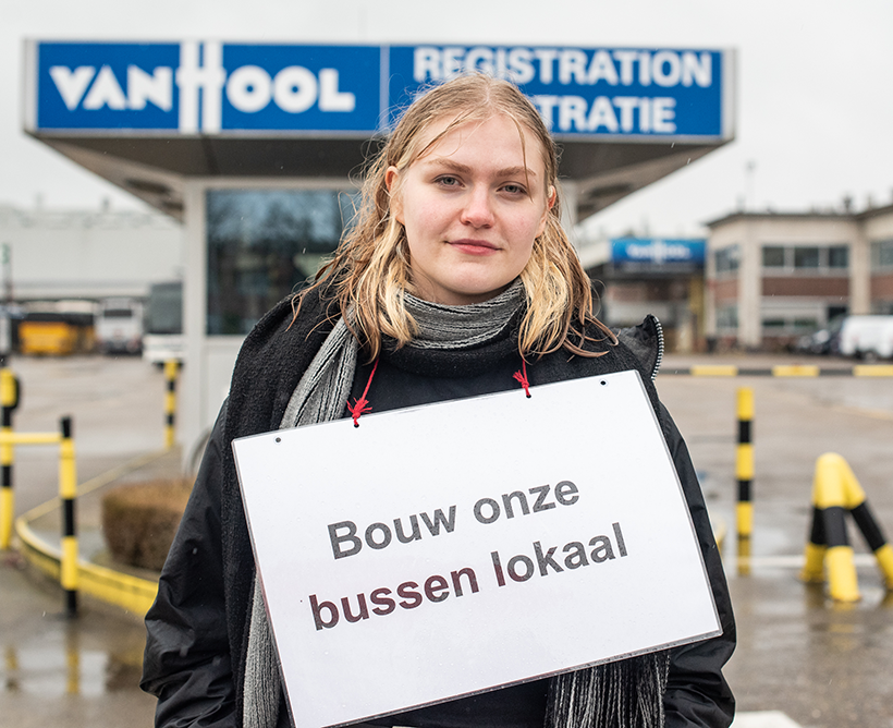 Een jonge vrouw staat voor de Van Hool fabriek met een bord waarop staat "Bouw onze bussen lokaal".