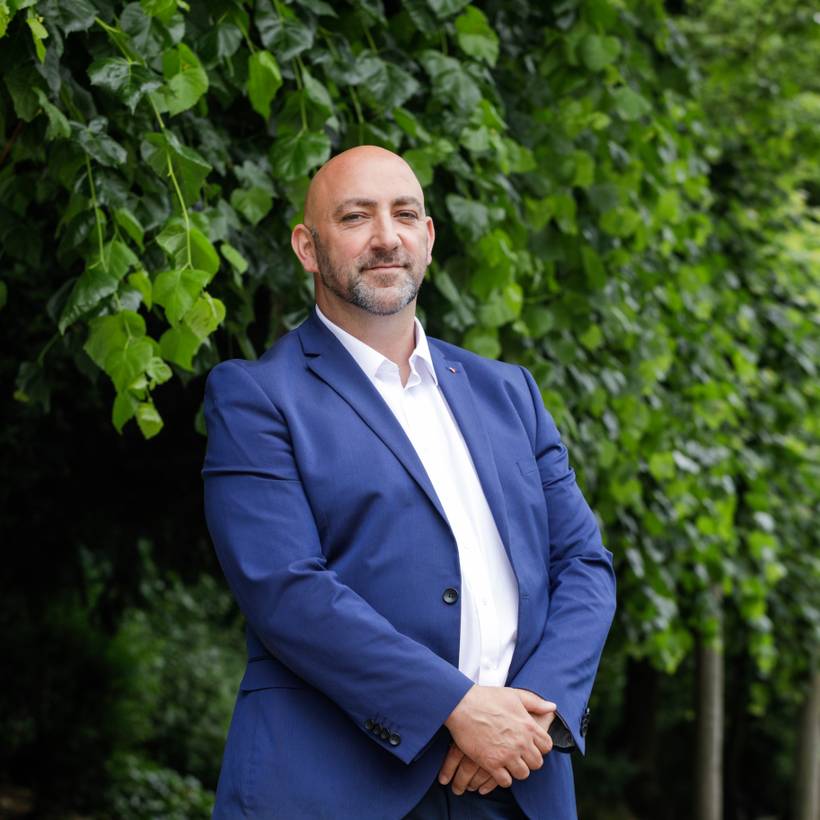 Gaby Colebunders se tient devant une rangée d'arbres. Il porte un blazer bleu foncé et une chemise blanche. Il tient ses mains devant son ventre et sourit avec précaution. 