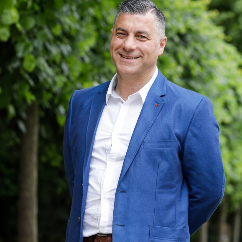 Roberto D'Amico se tient devant une rangée d'arbres. Il porte un blazer bleu foncé et une chemise blanche. Il a les mains derrière le dos. Il sourit amicalement.