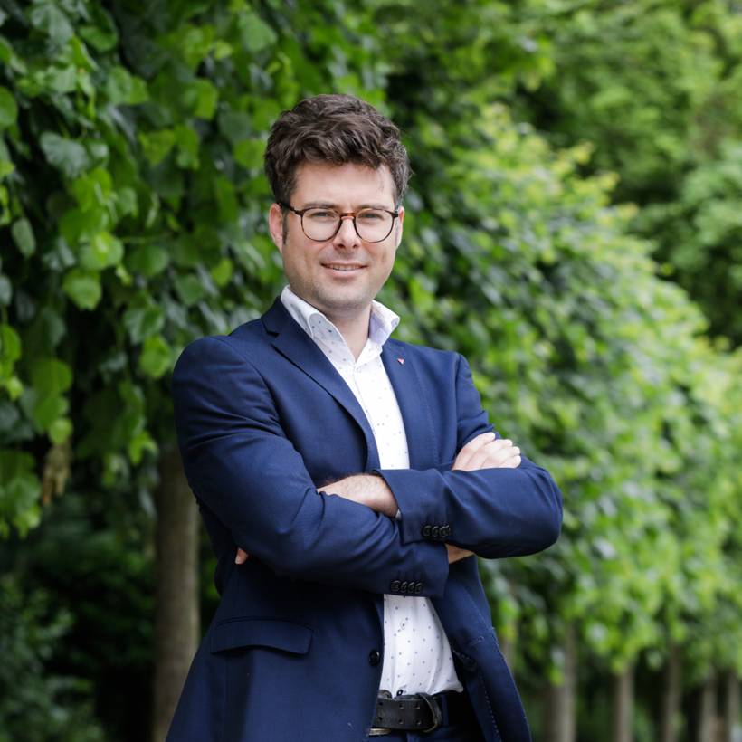 Steven De Vuyst se tient devant une rangée d'arbres. Il porte un blazer bleu foncé et une chemise blanche. Il sourit chaleureusement et a les bras croisés.