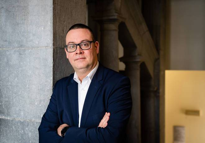 Raoul Hedebouw porte une veste sombre et une chemise blanche. Il est appuyé contre un mur, les bras croisés.