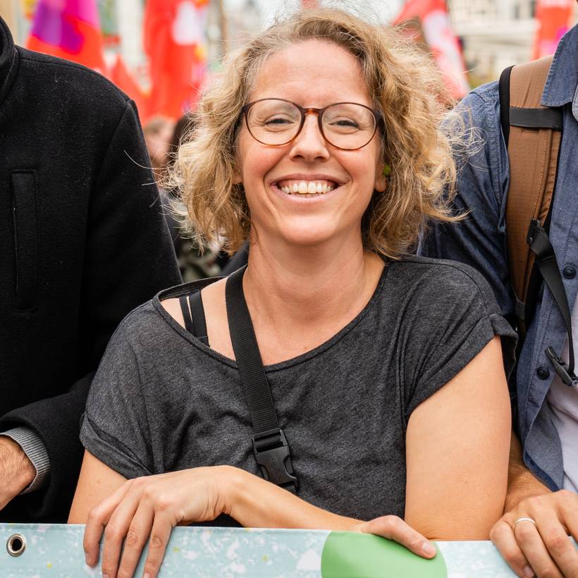 Greet Daems lors d'une manifestation sur le climat. Elle tient une banderole d'une main. Derrière elle, on peut voir des drapeaux du PVDA. Elle porte un T-shirt gris.