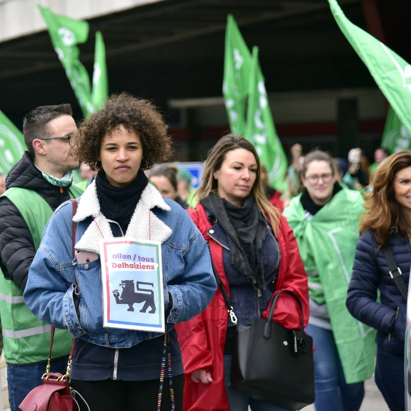Plusieurs personnes se tienne debout devant un Delhaize. Elles portent des chasubles et des drapeaux syndicaux verts et rouges. À l'avant-planc, une jeune femme brune vêtue d'une veste en jeans porte autour du coup l'affiche "Tous Delhaiziens" du PTB. 