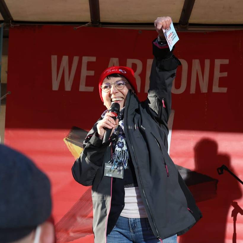 Nadia Moscufo tient un micro dans sa main et lève l'autre main en l'air comme un poing. Elle porte un chapeau rouge sur lequel on peut lire "Rebel with a Heart". Derrière elle, un drapeau rouge portant l'inscription "We Are One" est suspendu.