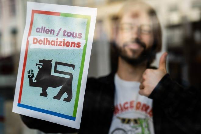 Un jeune homme placadre une affiche du PTB « Tous Delhaiziens » derrière une vitre. Il fait un pouce en l'air.