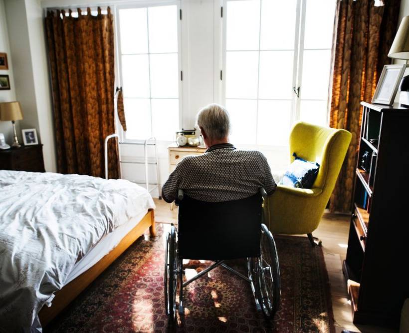 Een bejaarde man zit in een rolstoel, met zijn rug naar de camera. Hij zit in een kamer in een woonzorgcentrum.