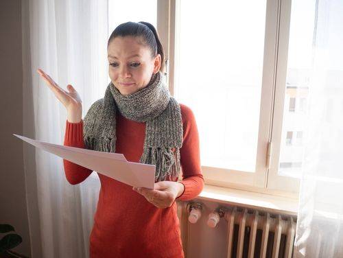 Een vrouw kijkt bezorgd naar een rekening. Ze draagt een grijze sjaal en een rood truitje. Ze staat met haar rug tegen de verwarming.