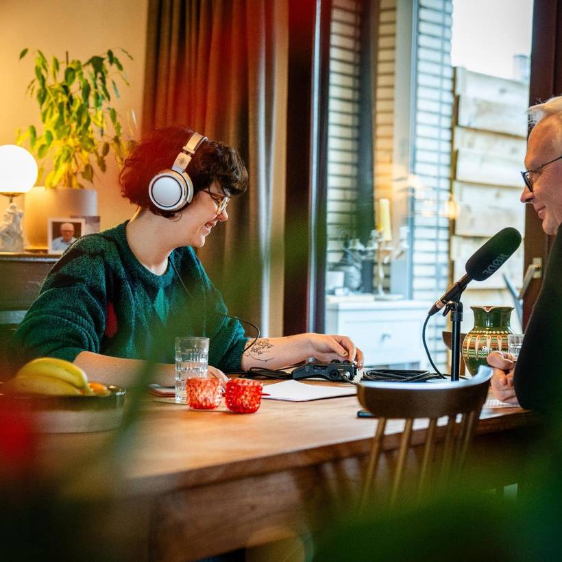 Lilo van Fakto zit aan tafel met Peter Mertens. Peter spreekt in een microfoon, terwijl Lilo luistert en op een opnameapparaat bijstelt.