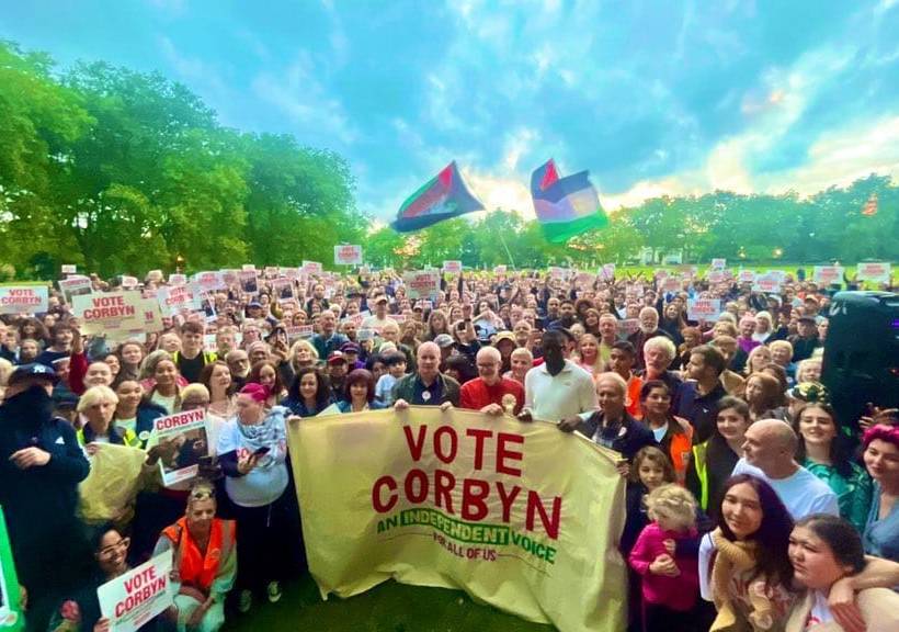 Jeremy Corbyn staat voor een menigte. Een vlag met "Vote Corbyn" hangt voor hen. 