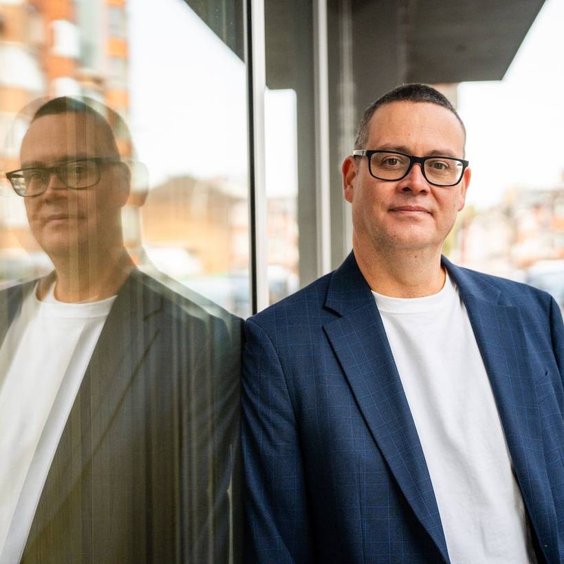 Raoul Hedebouw in de straten van Luik. Hij staat geleund tegen een venster en zijn weerspiegeling is goed zichtbaar.