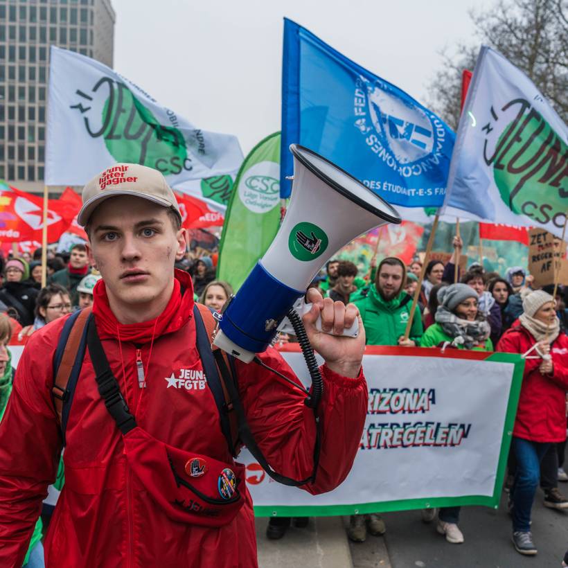 Een jonge man met een megafoon staat voor een spandoek en daarachter syndicalisten en vlaggen van alle vakbonden