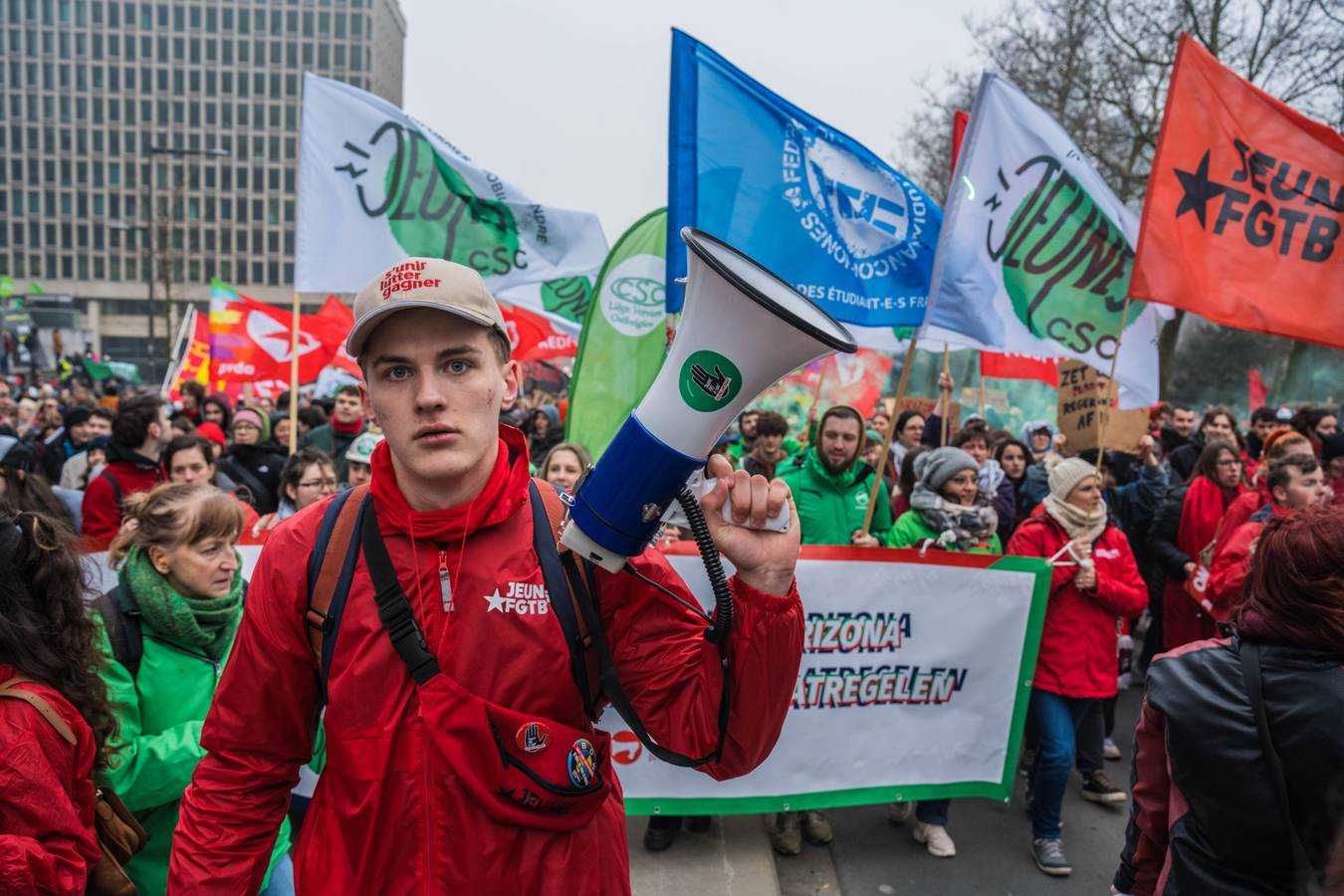 Een jonge man met een megafoon staat voor een spandoek en daarachter syndicalisten en vlaggen van alle vakbonden