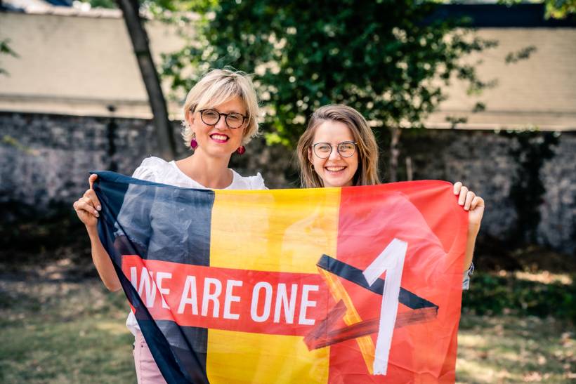 Sofie Merckx se tient debout dehors à côté de Pauline Boninsegna. Elles portent ensemble le drapeau belge où figure le slogan « We Are One »
