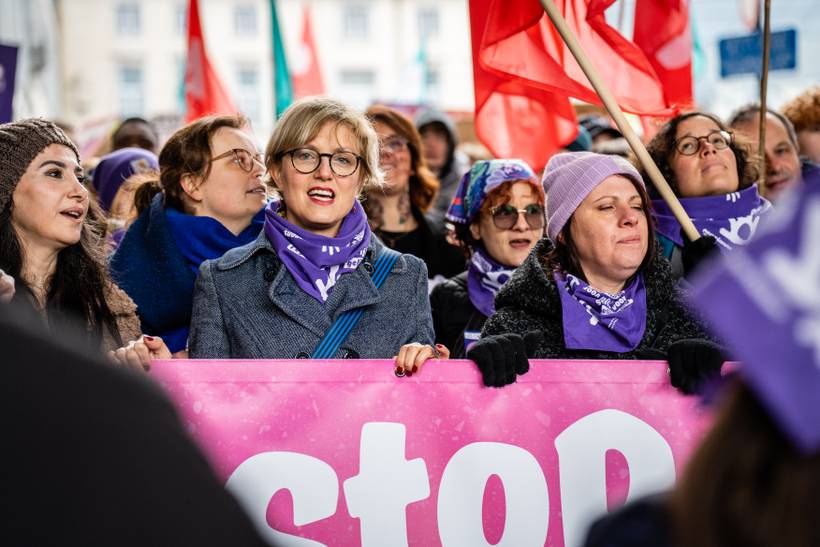 Sofie Merckx marche en tête de la manifestation contre les violences faites aux femmes. Elle tient d'une main une banderole sur laquelle on peut lire "Stop aux violences faites aux femmes". Elle porte un foulard mauve du mouvement de femmes Marianne.
