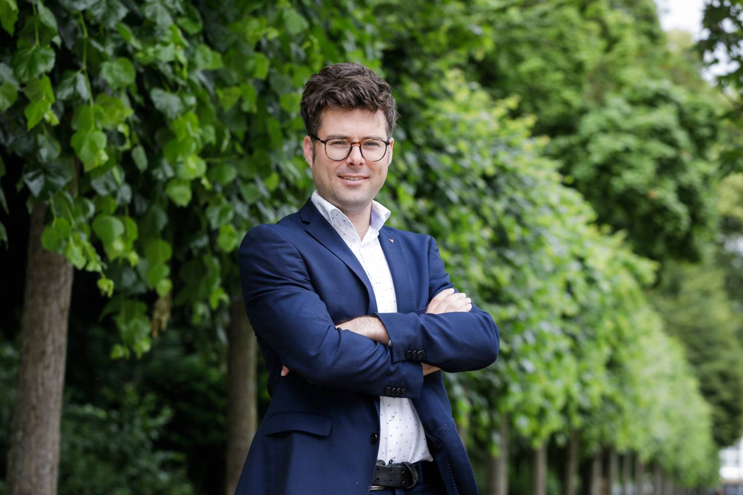 Steven De Vuyst se tient devant une rangée d'arbres. Il porte un blazer bleu foncé et une chemise blanche. Il sourit chaleureusement et a les bras croisés.