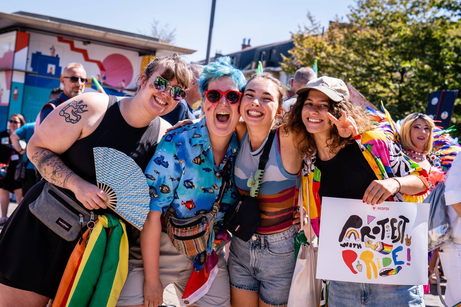 Vier mensen op de Pride-betoging. Ze dragen kleurrijke kleren. 1 iemand heeft een poster vast van RedFox, waarop staat "Protest with Pride".