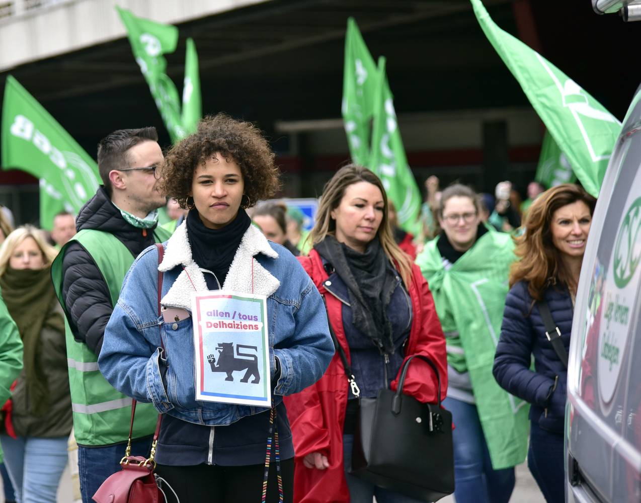 Plusieurs personnes se tienne debout devant un Delhaize. Elles portent des chasubles et des drapeaux syndicaux verts et rouges. À l'avant-planc, une jeune femme brune vêtue d'une veste en jeans porte autour du coup l'affiche "Tous Delhaiziens" du PTB. 