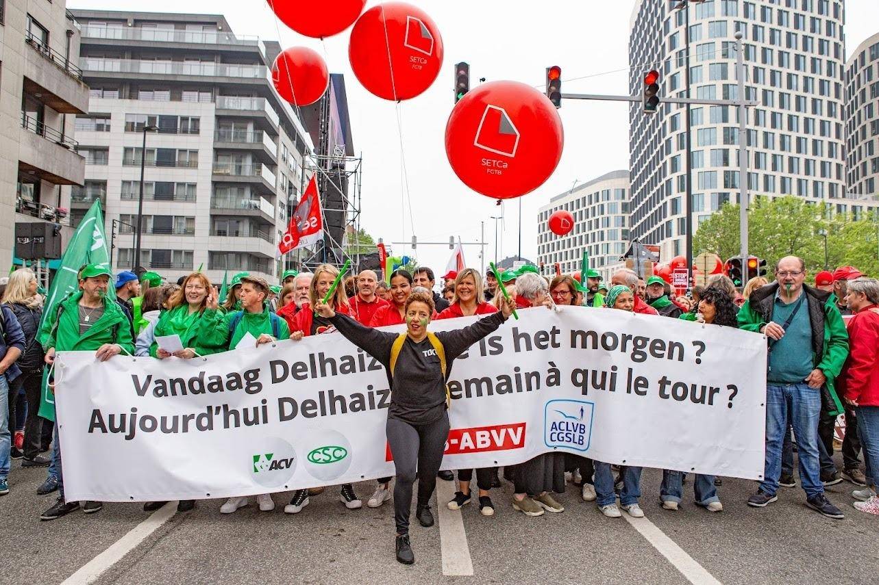 Vakbondsbetoging in Brussel. Een spandoek met daarop: "Vandaag Delhaize, aan wie is het morgen?" Voor het spandoek staat een persoon met de handen in de lucht en percussiestokken in de handen. Achter de spandoek staan vakbondsleden van het ACV en het ABVV