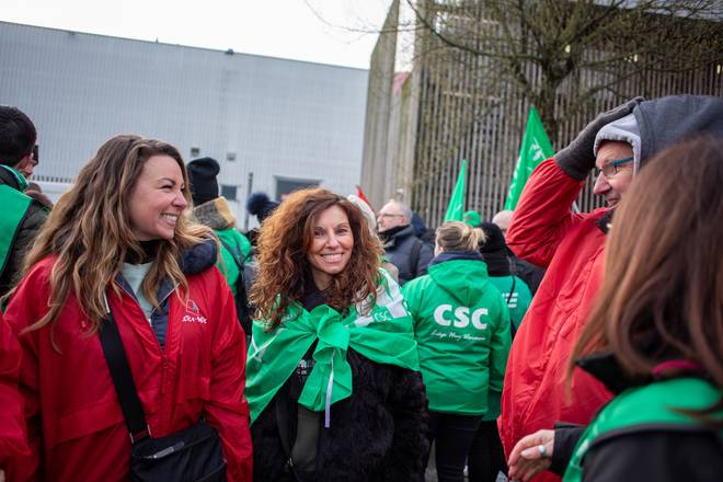 Deux travailleuses de Delhaize se trouvent au milieu d'une manifestation syndicale à Bruxelles. L'une porte une veste rouge FGTB, l'autre verte CSC. Elles sont très souriantes. Autour d'elles, il y a beaucoup de syndicalistes qui participent.