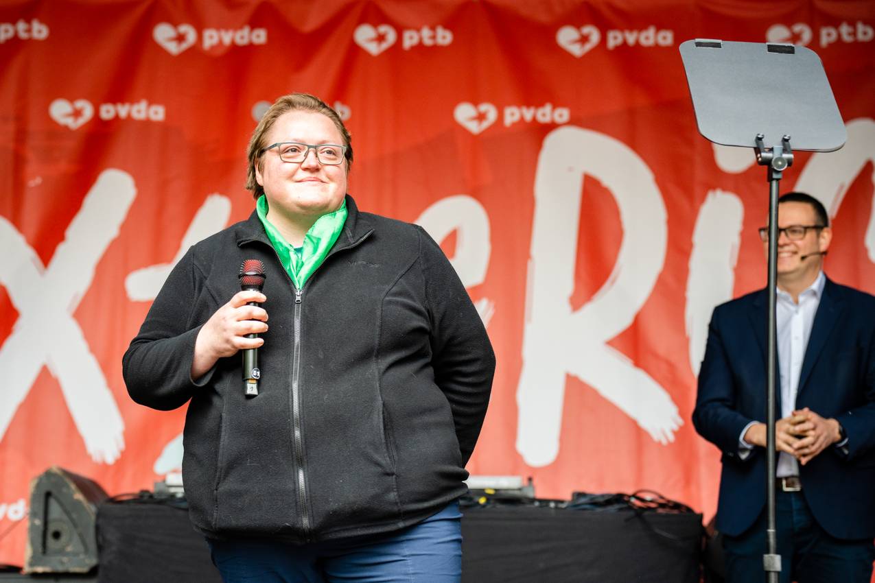 Agnès staat voor de "Tax The Rich" banner van de PVDA. Ze heeft een microfoon in haar hand. Ze draagt een groen sjaaltje om de nek en een zwarte jas. Ze heeft een ontroerde blik.